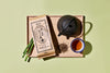 Tea set with a teapot, tea leaves, and a tea package on a wooden tray against a light green background.